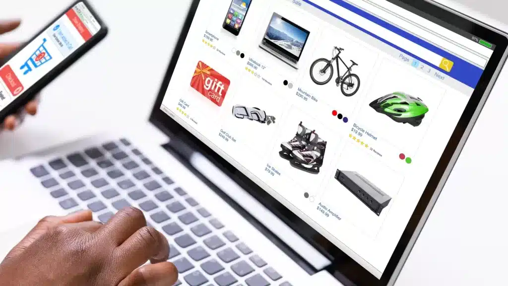 On the white desktop of the laptop, the screen displays "Shop Online", the right hand is on the keyboard, and the left hand clicks the shopping cart icon on the phone.
